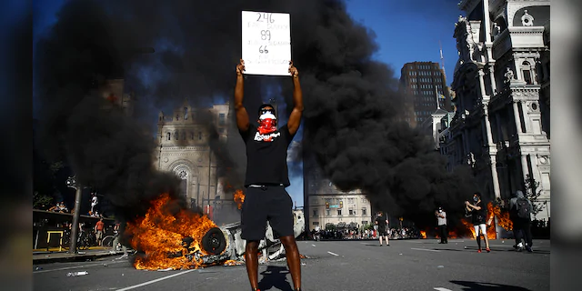 A vehicle is on fire behind a protester holding a sign during a rally on Saturday, May 30, 2020, in Philadelphia, over the death of George Floyd, a black man who died after being taken into police custody in Minneapolis. (AP Photo/Matt Rourke)