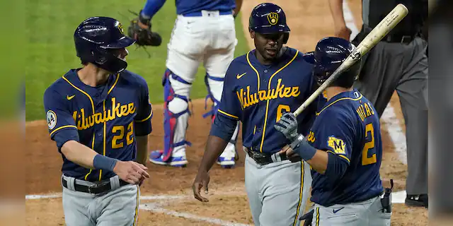 Milwaukee Brewers' Christian Yelich (22), Lorenzo Cain (6) and Luis Urias (2) celebrate after Yelich and Cain scored on an Orlando Arcia double in the sixth inning of a preseason baseball game against the Texas Rangers in Arlington, Texas, Monday, March 29, 2021. (AP Photo/Tony Gutierrez)