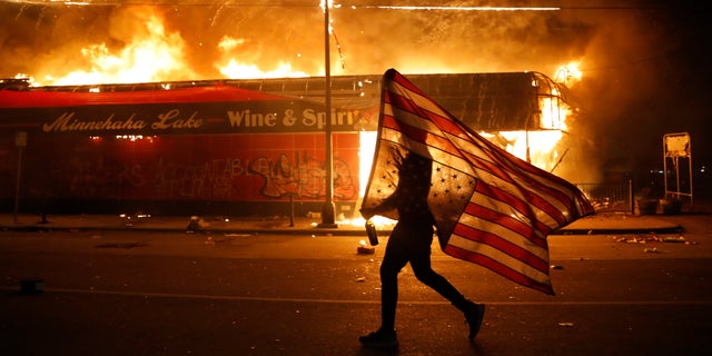 FILE - A protester carries a U.S. flag upside down, a sign of distress, next to a burning building, Thursday, May 28, 2020, in Minneapolis during protests over the death of George Floyd. Speaking at the Republican National Convention, President Donald Trump said, "The Republican Party condemns the rioting, looting, arson and violence we have seen in Democrat-run cities all, like Kenosha, Minneapolis, Portland, Chicago and New York, and many others." (AP Photo/Julio Cortez, File)