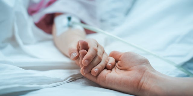 A child in a hospital bed holds their parents hand.