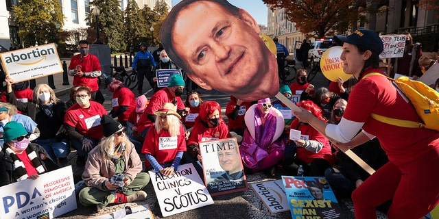 An activist with The Center for Popular Democracy Action holds a photo of Justice Samuel Alito as they block an intersection in front of the Supreme Court on Dec. 1, 2021, in Washington.
