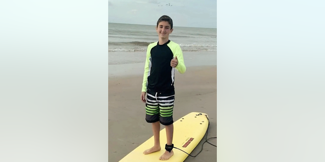 Nate Bronstein stands on a surfboard and gives a thumbs up.