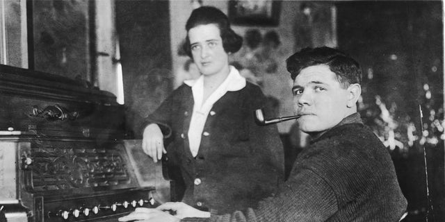 American baseball player Babe Ruth (1895-1948) — born George Herman Ruth Jr. — smokes a pipe as he plays a piano while his wife, Helen Ruth (nee Woodford, 1897-1929) stands next to him in the 1910s.