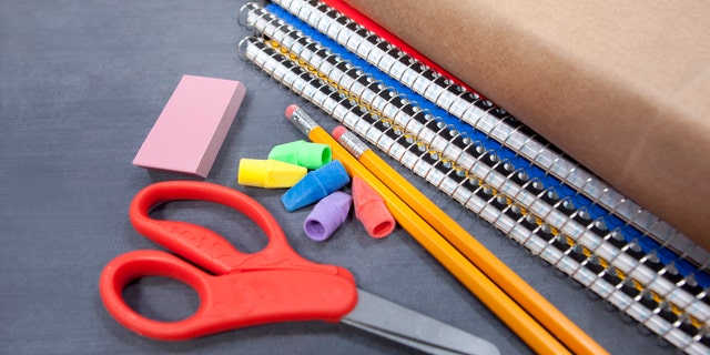 A pair of safety scissors is seen near pencils, erasers and spiral notebooks. Small children typically use safety scissors to practice cutting safely.