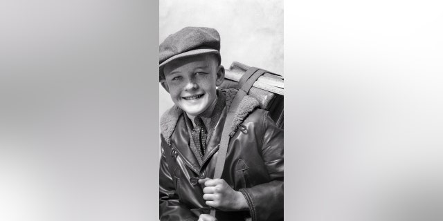 A smiling boy wears a wool newsboy cap, a leather bomber jacket and a necktie while holding a book strap. 