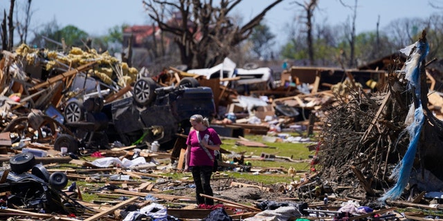 A woman walks near an uprooted tree, a flipped vehicle and debris from homes damaged by a tornado on March 27, 2023, in Rolling Fork, Miss.