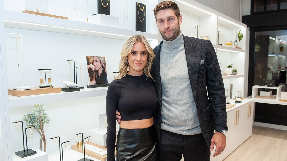 Kristin Cavallari in a long sleeve black crop top shirt and leather bottoms smiles next to Jay Cutler in a black suit jacket and grey turtleneck