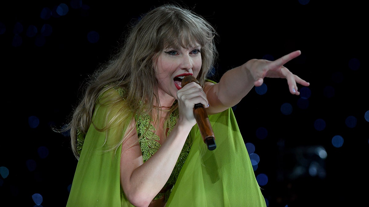 Taylor Swift in a green dress reaches her hand out on stage