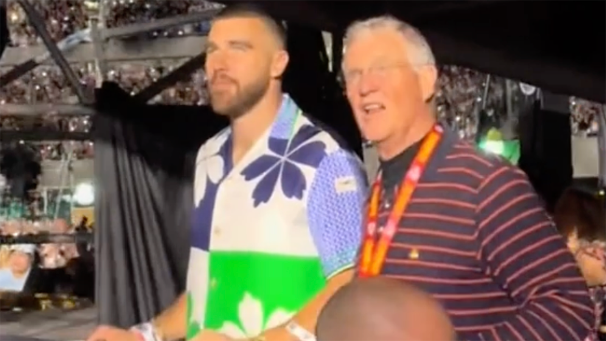 Travis Kelce in a patterned shirt stands next to Scott Swift in a collared shirt and Kansas City Chiefs lanyard