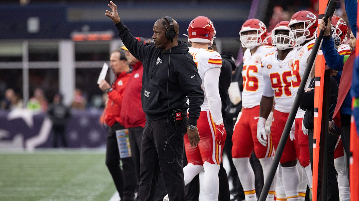 Dave Merritt in all black wearing a headset with his arm up in the air as Kansas City Chiefs players stand behind him