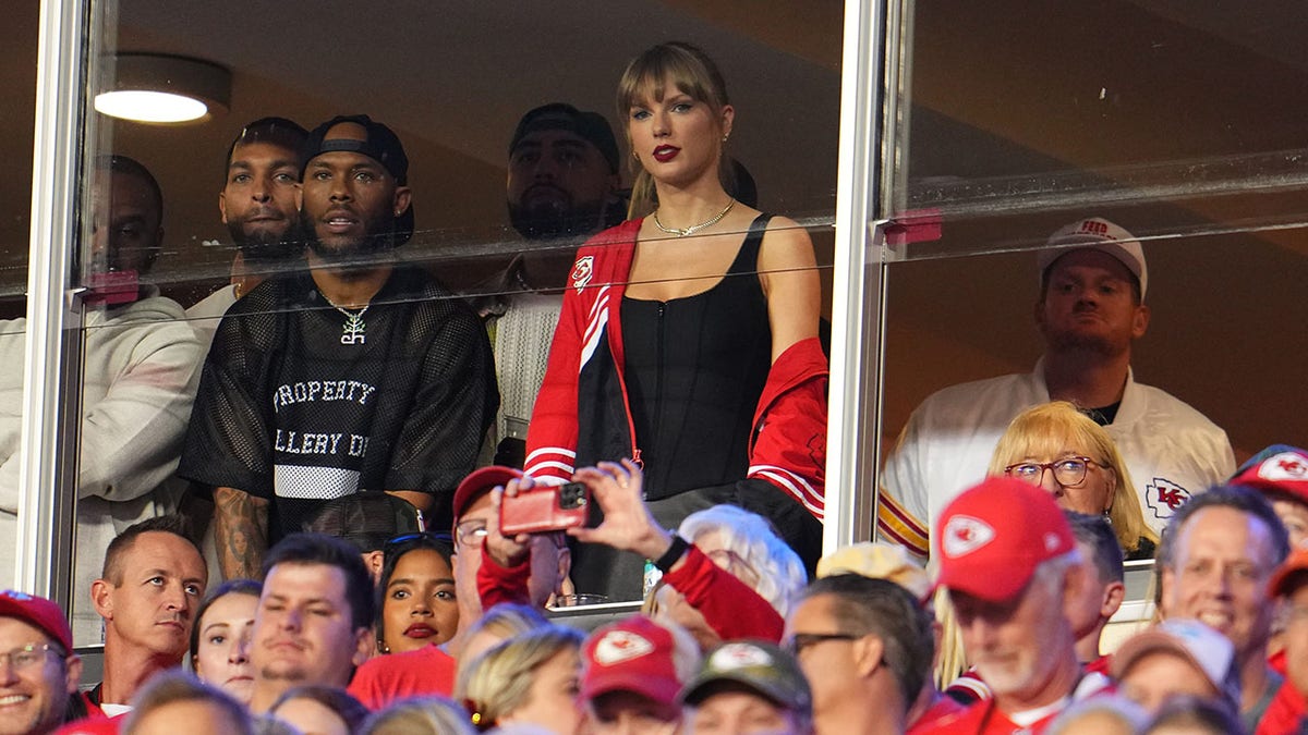 Taylor Swift in a black tank top and red jacket stands in the suite watching the Chiefs play surrounded by people