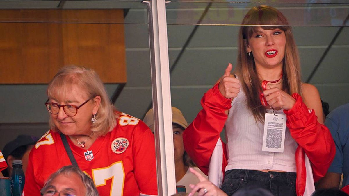 Taylor Swift in a white tank top and red jacket makes a thumbs up as she stands in a suite next to Donna Kelce in a Travis Kelce Chiefs jersey