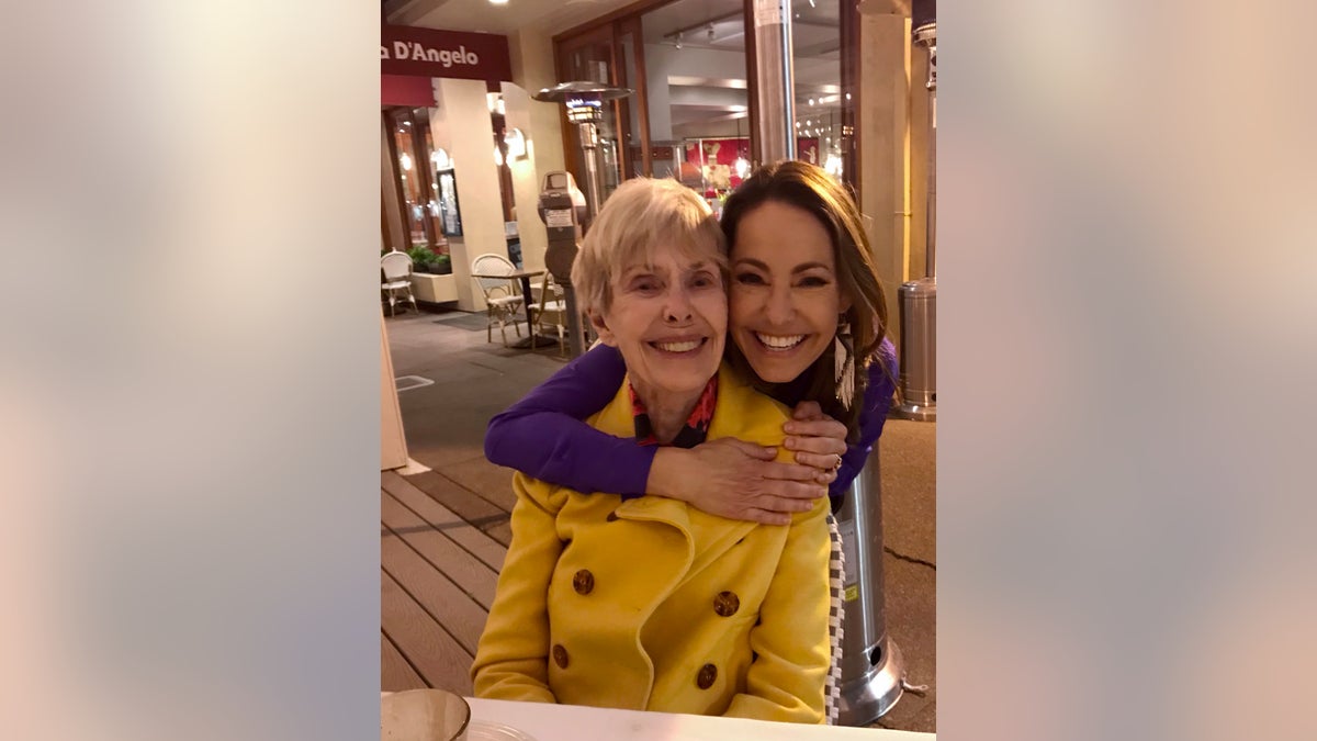 Barbara Rush in a yellow jacket being hugged from behind by her daughter as they are both smiling
